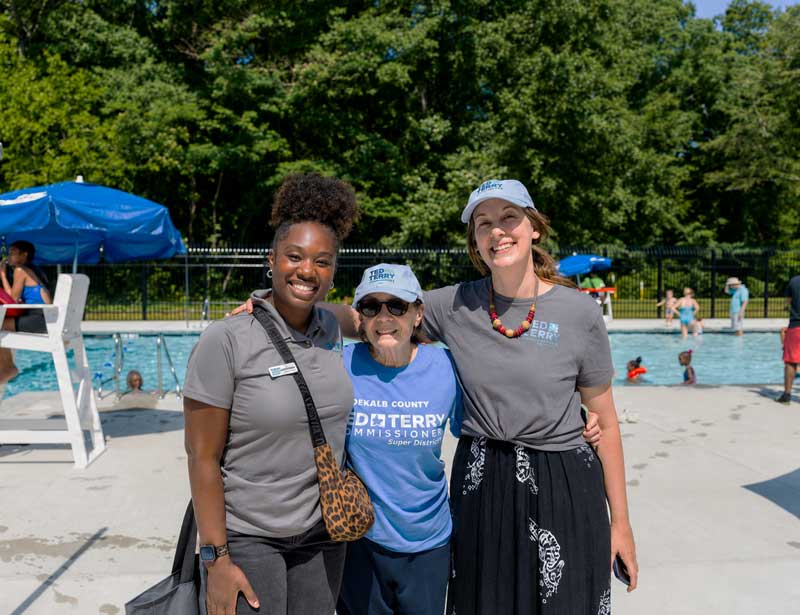 Three smiling women pose by a swimming pool, one wearing a "Dekalb County Commissioner" shirt.