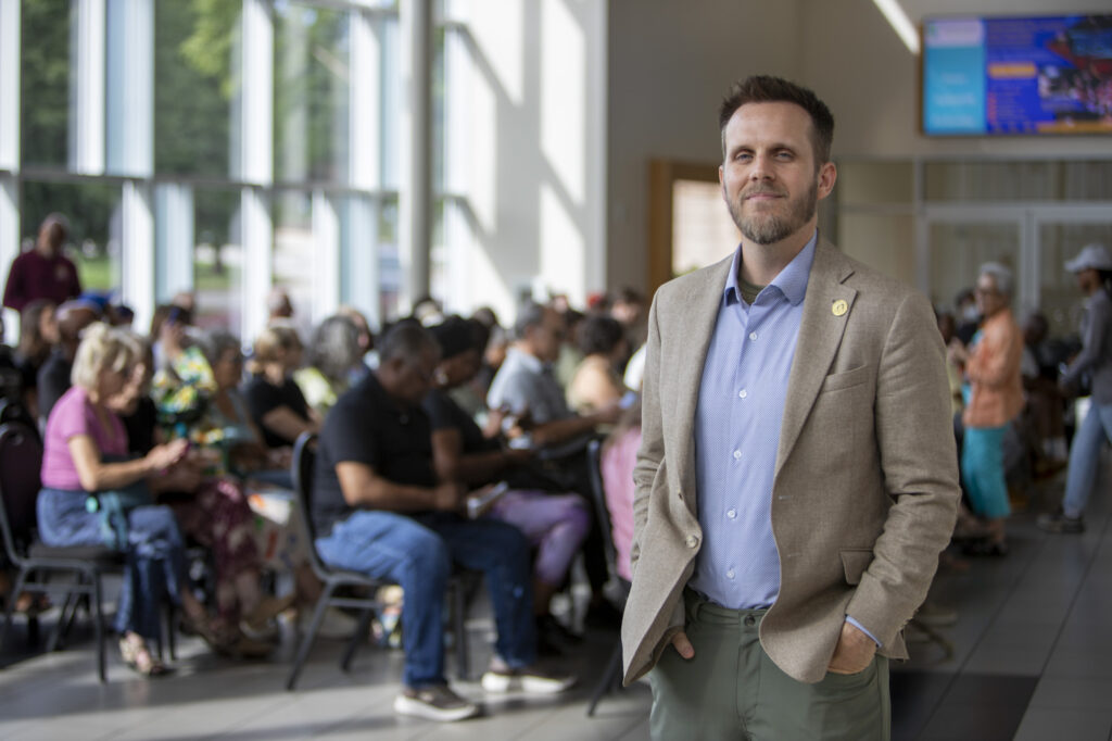 Man in a beige blazer stands confidently in a busy, sunlit waiting area with seated and standing people behind him.