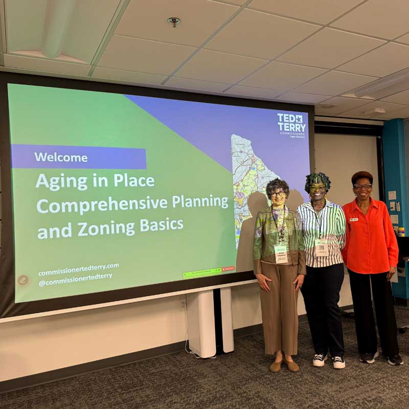 Three people stand in front of a presentation screen about "Aging in Place Comprehensive Planning" with a map.