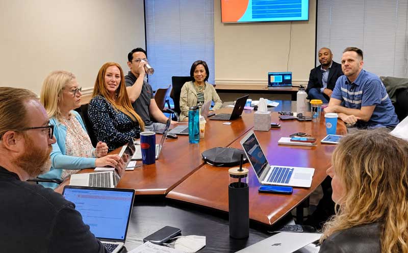 Diverse team in a conference room engaging in a meeting with laptops open and a presentation on the wall.