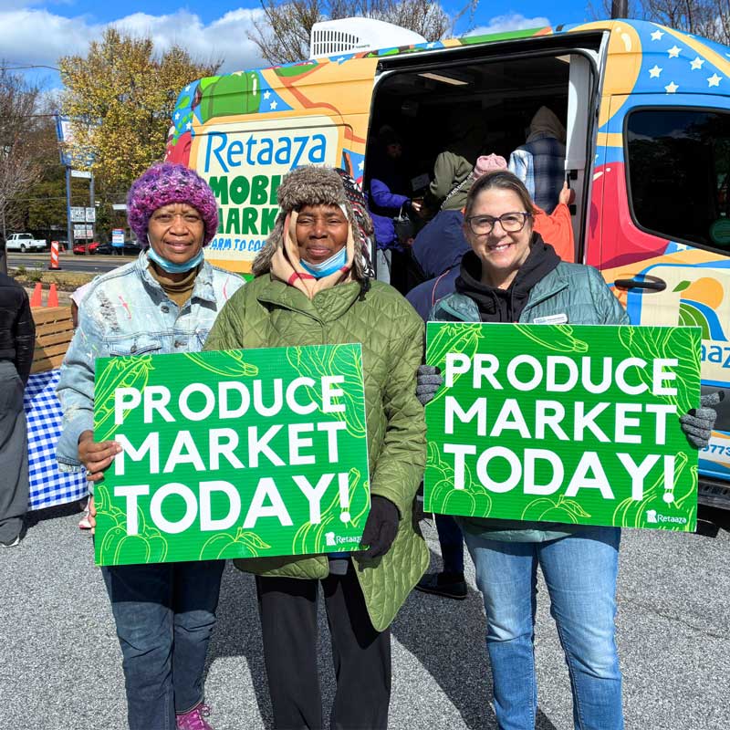 Three people holding "Produce Market Today" signs in front of a colorful mobile market truck.