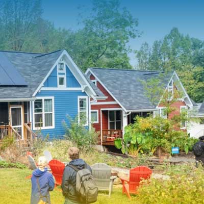 Colorful houses with solar panels in a lush setting, children walking past red chairs and greenery in the foreground.