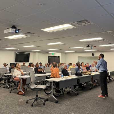 A lecturer addresses an attentive audience in a modern conference room with multiple screens and projectors.