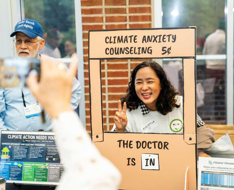 Woman offering "Climate Anxiety Counseling" smiles, giving a peace sign, next to a man wearing a "Make Earth Cool Again" cap.