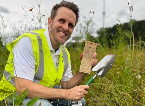 Man in a neon vest kneeling in a field, holding a spade and a wooden box, surrounded by tall grass.