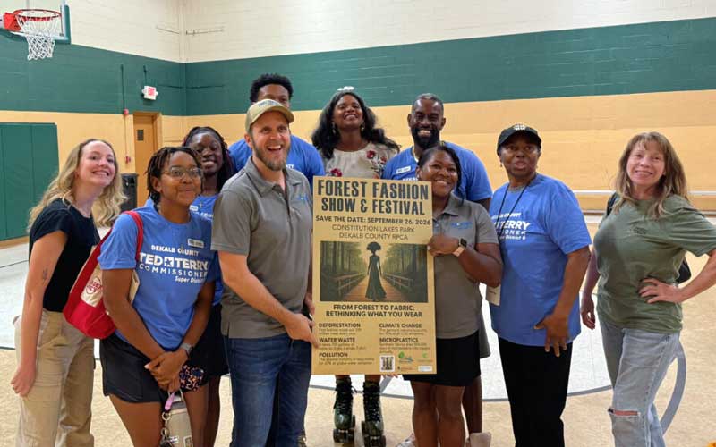 Group of people smiling and holding a "Forest Fashion Show & Festival" poster in a gymnasium.