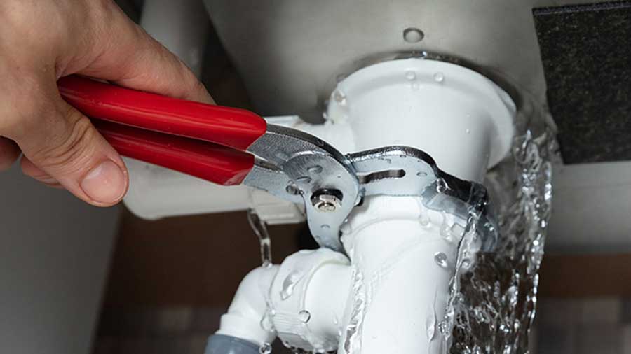 Hand using wrench to fix leaking pipe under sink with flowing water.