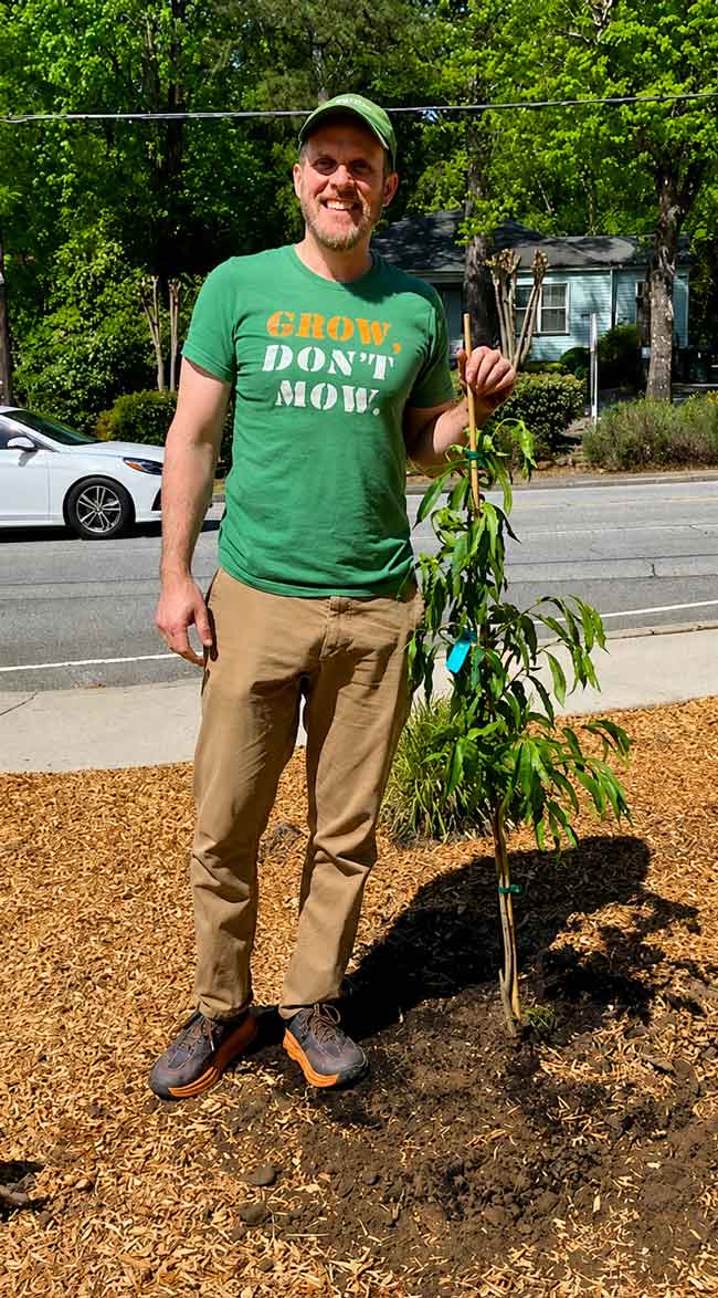 Man smiling next to a young tree with a "Grow Don't Mow" shirt, standing on a mulched area.