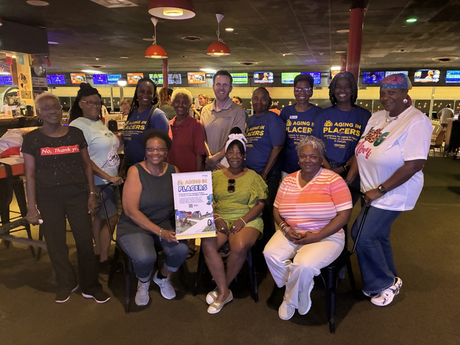 Group of people at an event holding a poster with "Aging in Placers" text, in a room with colorful lights and screens.