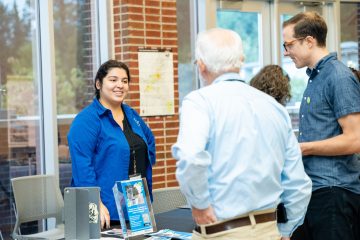 A woman in a blue shirt engages with two attendees at a registration table in a bright, spacious indoor setting.