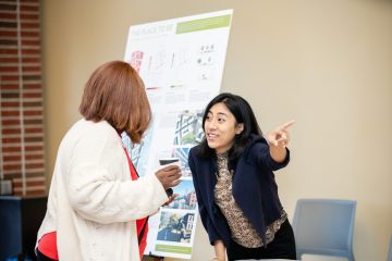 Woman in discussion, gesturing towards a presentation board featuring architectural designs.