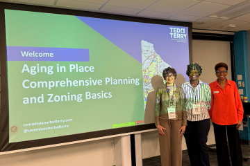 Three individuals stand in front of a presentation screen titled "Aging in Place Comprehensive Planning and Zoning Basics".