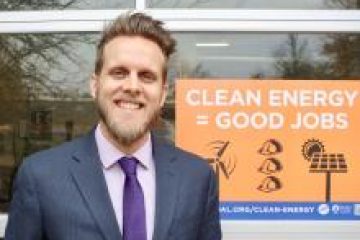 Smiling man in a suit stands beside a clean energy sign reading "Clean Energy = Good Jobs" near a window.