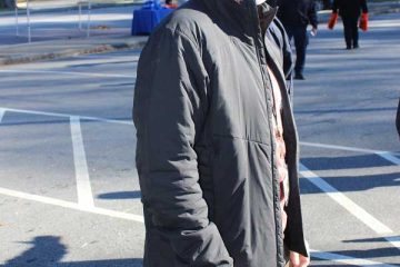 Person in a black jacket and white mask stands outside near orange canopy, trees in the background.