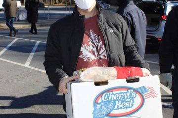 Man in mask holds a "Cheryl's Best" box at outdoor event, surrounded by people and tents in a park setting.