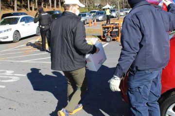Two men in a parking lot, one carrying a box, near a line of parked cars and police in the background.