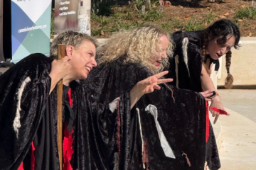 Three women in black cloaks performing theatrically outdoors, gesturing dramatically with an engaged audience.