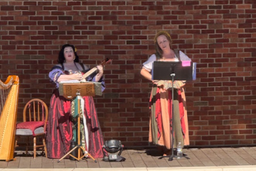 Two women in period costumes perform with a guitar and sheet music stand against a brick wall.