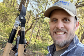 Man smiling while holding garden loppers, standing in a forested area wearing a cap and jacket.